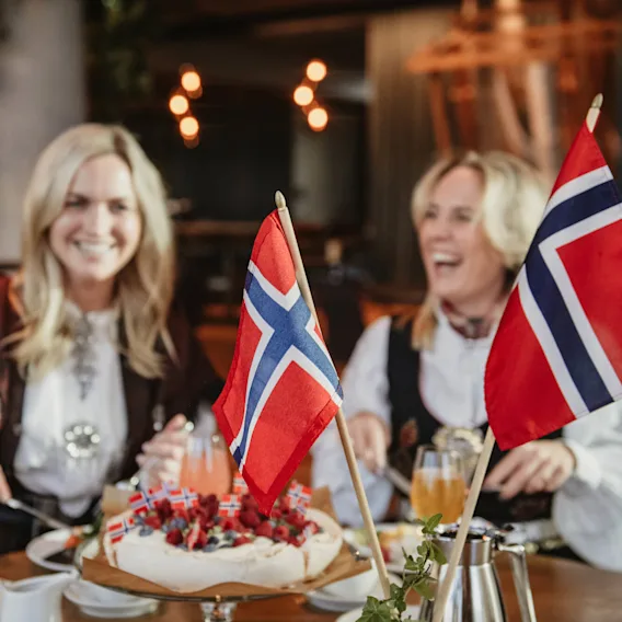 To kvinner i bunad ler og skjærer i en kake dekorert med norske flagg, i en koselig restaurantinnstilling.