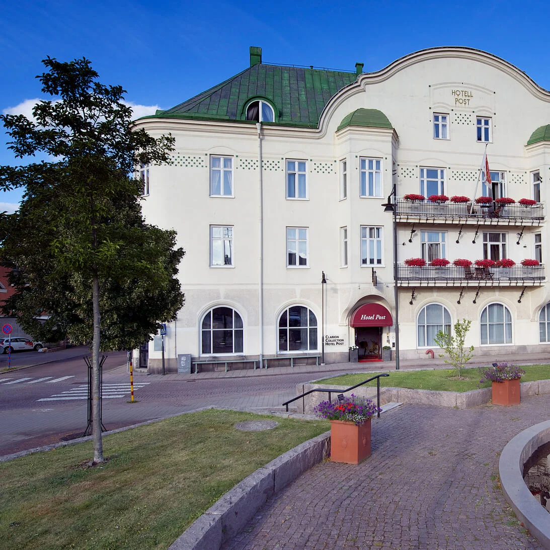 A three-story building with "HOTELL POST" sign and balconies, on a sunny day with clear skies, beside a small pond and park benches.
