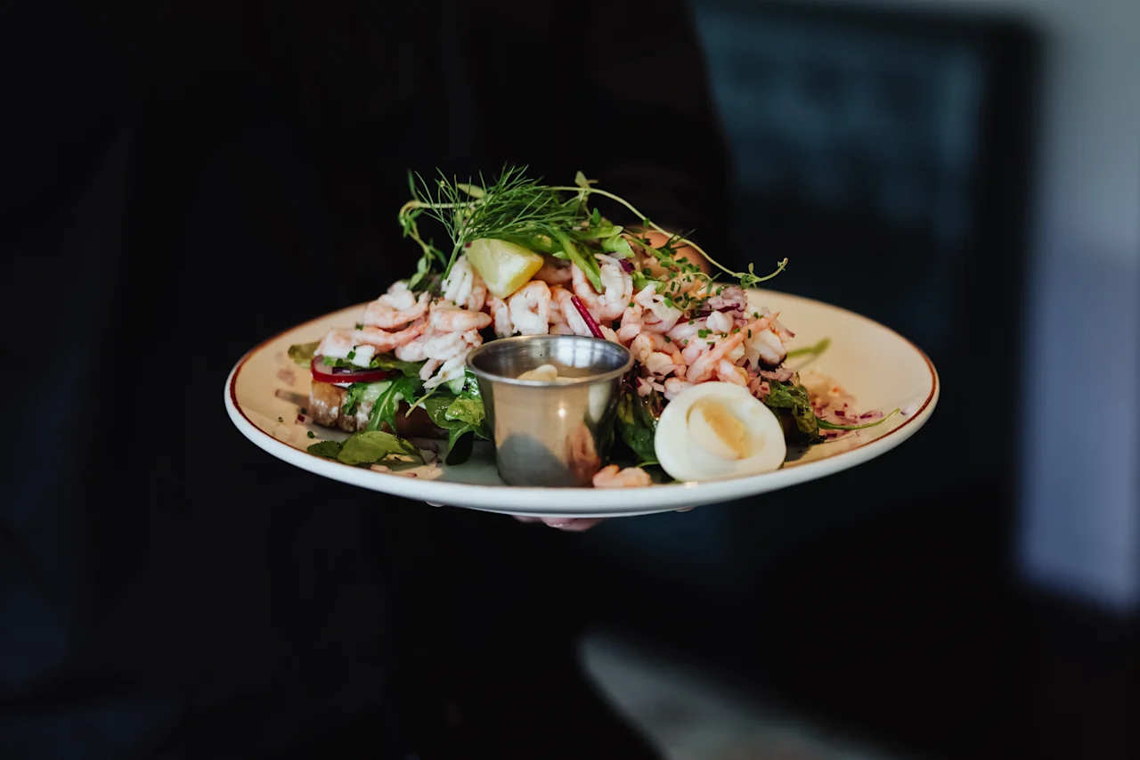 A plate of shrimp salad with lemon wedges, fresh greens, and a side of dressing is held by a person in a dimly lit restaurant.