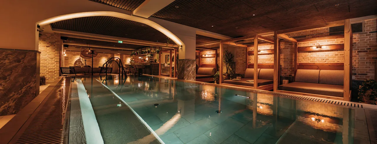 A rectangular indoor pool with gentle lighting, surrounded by lounge seating and wooden beams, offers a warm and inviting atmosphere for relaxation.