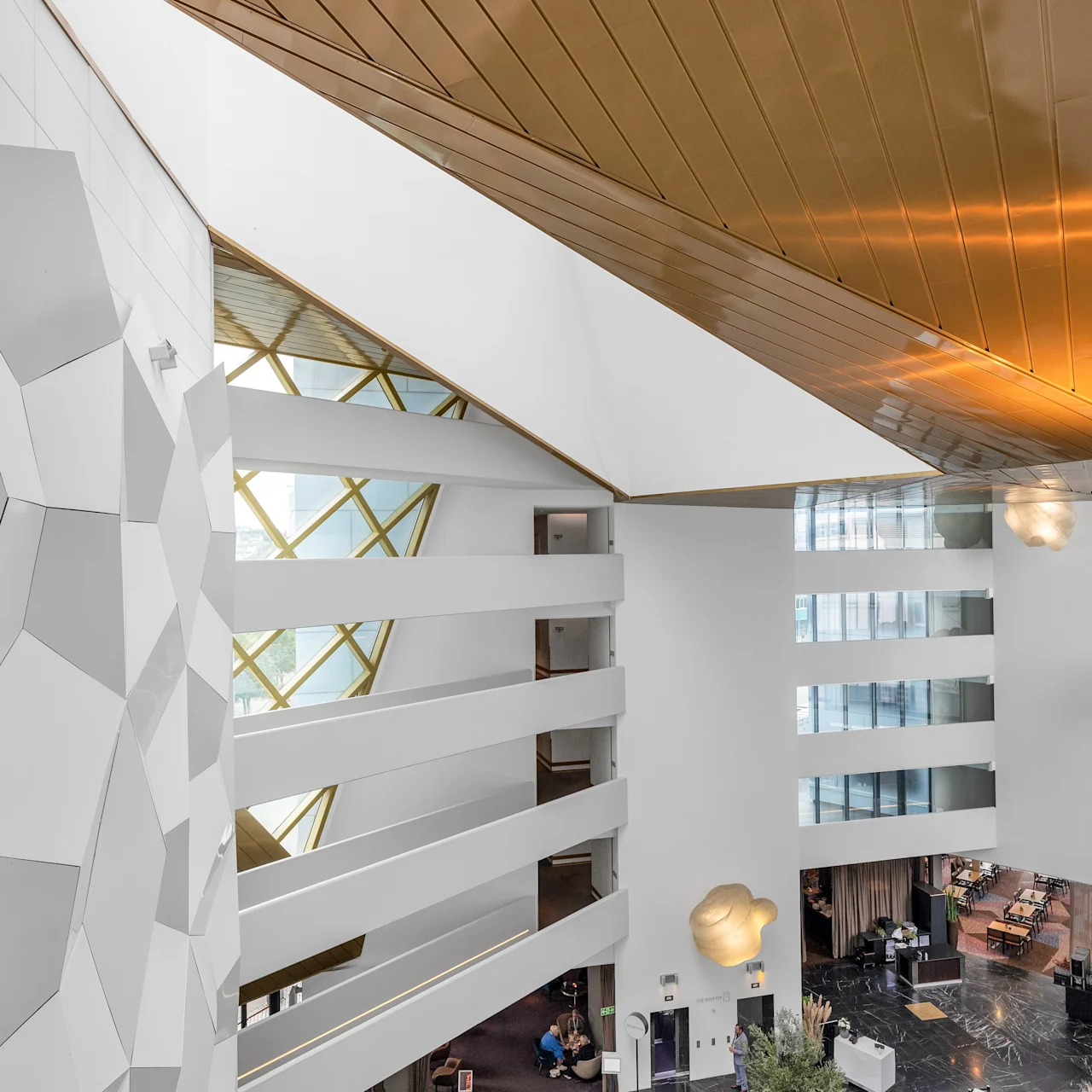 A large, modern atrium features geometric white walls and balconies overlooking a lobby. Natural light streams through upper windows, gold ceiling panels reflect light, creating an elegant and open space.