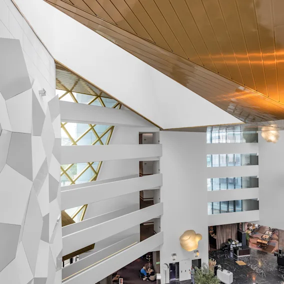 Et stort, moderne atrium har geometriske hvite vegger og balkonger med utsikt over en lobby. Naturlig lys strømmer gjennom øvre vinduer, gulltakpaneler reflekterer lys, og skaper en elegant og åpen atmosfære.
