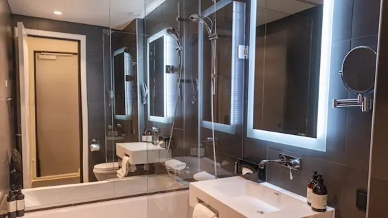 Moderne baderom med rektangulær vask, opplyst speil og glassdusj. Rommet har minimalistiske beslag og mørke fliser, noe som skaper en elegant og moderne atmosfære.