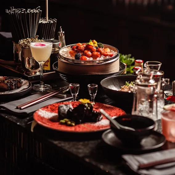 En mängd olika rätter och drycker är arrangerade på ett svagt upplyst restaurangbord, med frukt på is, en cocktail och små tallrikar med garneringar, omgivna av bestick och glas.