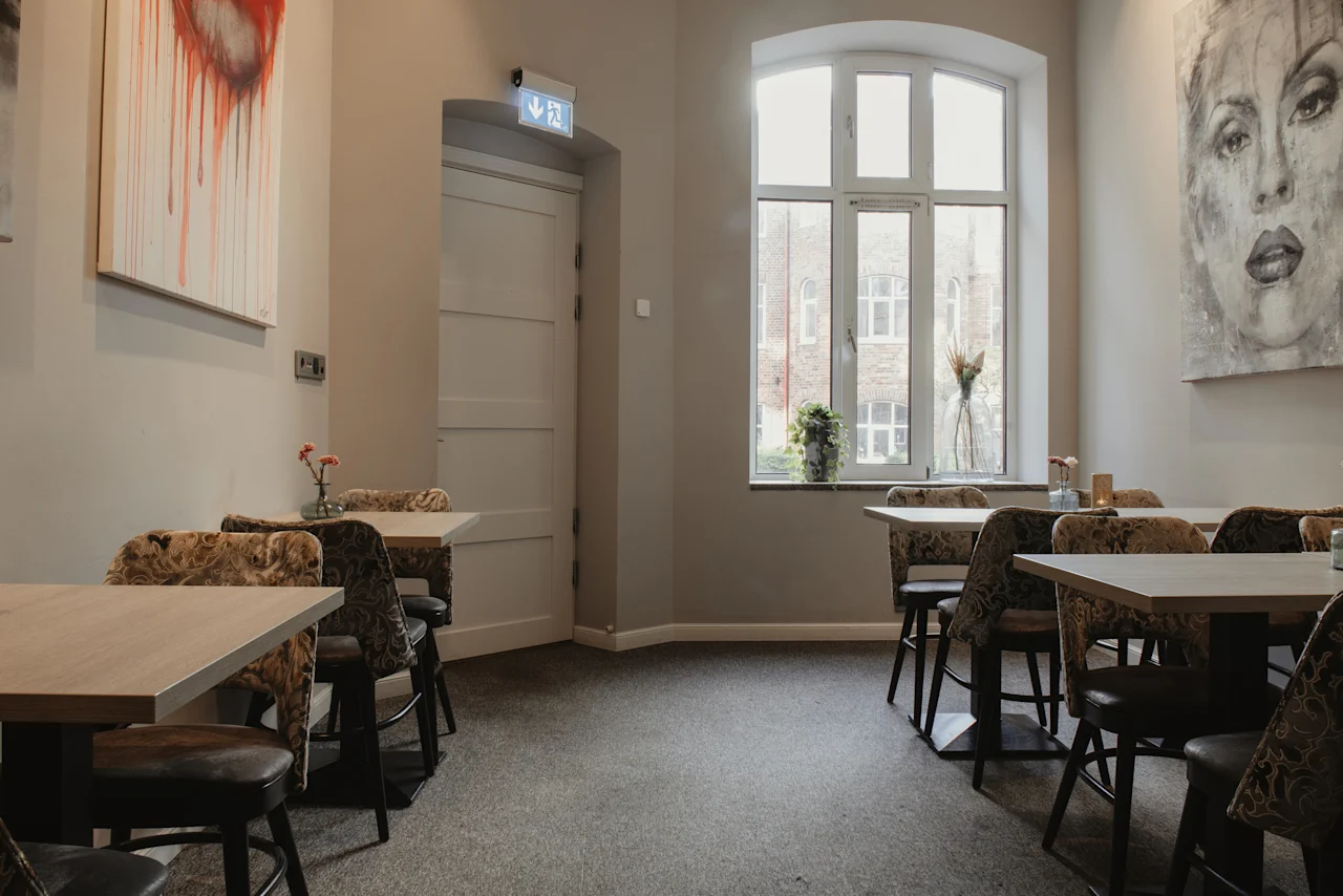 Et restaurantområde med borde, stole og kunstværker. Et stort vindue har udsigt til bygninger, og et udgangsskilt er over en dør.