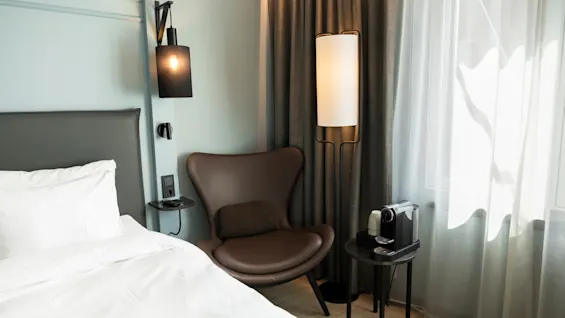 Et koselig hotellrom med en seng, en komfortabel stol og en kaffemaskin, opplyst av naturlig lys og stilige lamper.