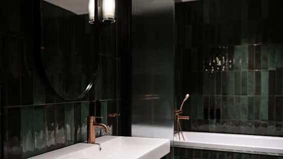 Vitt handfat med kopparblandare står oanvänt i ett modernt badrum. Mörkgröna kaklade väggar omger och reflekterar ljus från en närliggande lampa, vilket framhäver den eleganta, minimalistiska designen.