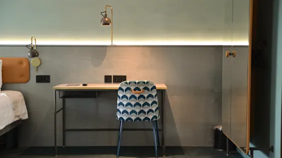 En mønstret stol står foran et minimalistisk skrivebord, under to lamper, mod en lys væg. En seng er delvist synlig til venstre.