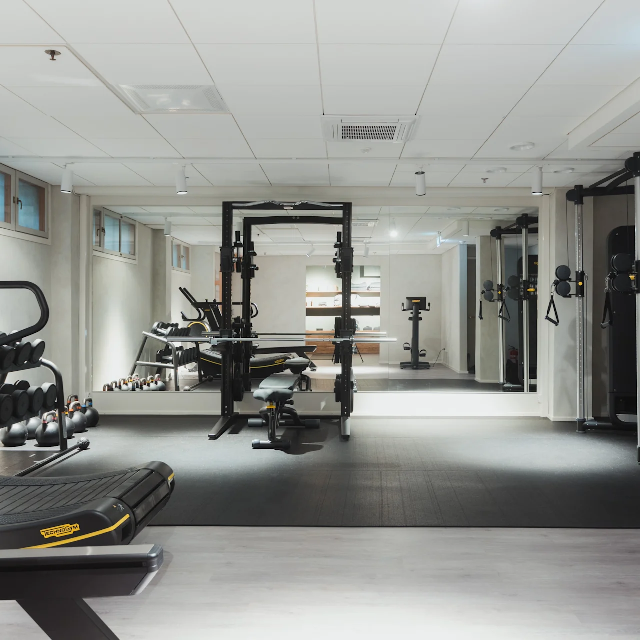 Løbebånd står i et rummeligt fitnesscenter, omgivet af vægte og træningsudstyr, med spejle der reflekterer rummet. Nødbelysning synlig i loftet.
