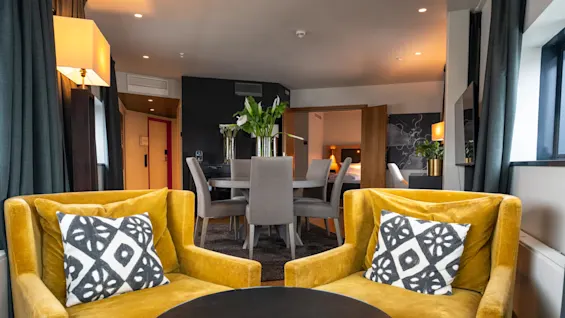 To sennepsfarvede lænestole med mønstrede puder vender mod et rundt sort bord i en moderne stue, der har et spiseområde med grå stole og vaser med blomster.