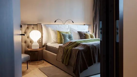 A neatly made bed with multiple pillows and a throw blanket, illuminated by a soft, glowing lamp on a bedside table, in a cozy, warmly lit bedroom.