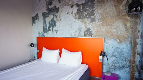 En pænt redt seng med hvide puder og lagner står mod et orange hovedgærde; en slidt, kunstnerisk væg og et natbord med lamper fuldender det moderne soveværelse.
