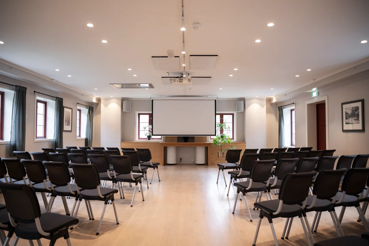 Konferanserom med rader av tomme stoler vendt mot en stor projektorskjerm. Rommet er godt opplyst med taklys og har innrammede bilder og vinduer i en moderne setting.
