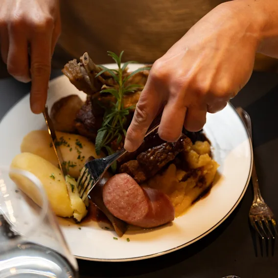 Hender bruker kniv og gaffel for å skjære i et solid måltid med kjøtt, poteter og pølse på en hvit tallerken.