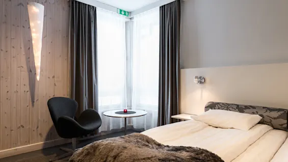Et moderne hotelværelse har en pænt redt seng, et lille rundt bord med et rødt lys og en sort stol. En oplyst vægdekoration og store vinduer med mørke gardiner giver en hyggelig atmosfære. Et nødudgangsskilt ses over vinduet.