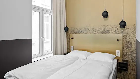 En pent senger med hvitt sengetøy hviler under en gul vegg med graffiti, i et moderne rom dekorert med to hengende svarte lamper og store vinduer med beige gardiner.