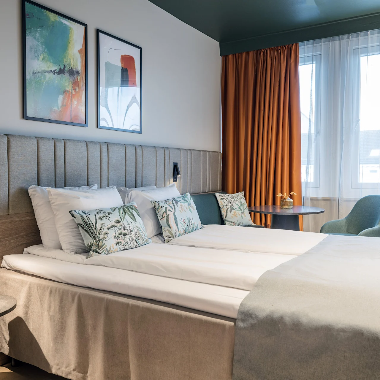 Siisti hotellivuode kukkatyynyineen on huoneessa, jossa on abstrakteja maalauksia seinällä. Lähellä oranssi verho peittää ikkunan, valaisten tuolia ja sivupöytää.