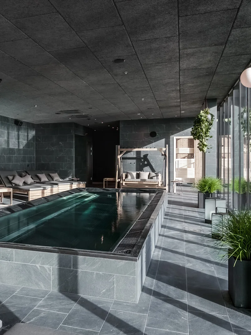 Suorakulmainen sisäuima-allas, jossa on kirkasta vettä, ympärillä pehmustettuja puukalusteita, modernissa kivihuoneessa, jossa suuret ikkunat ja vihreitä kasveja.