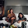Two people laughing and enjoying a relaxed breakfast in bed with drinks and food on a tray.