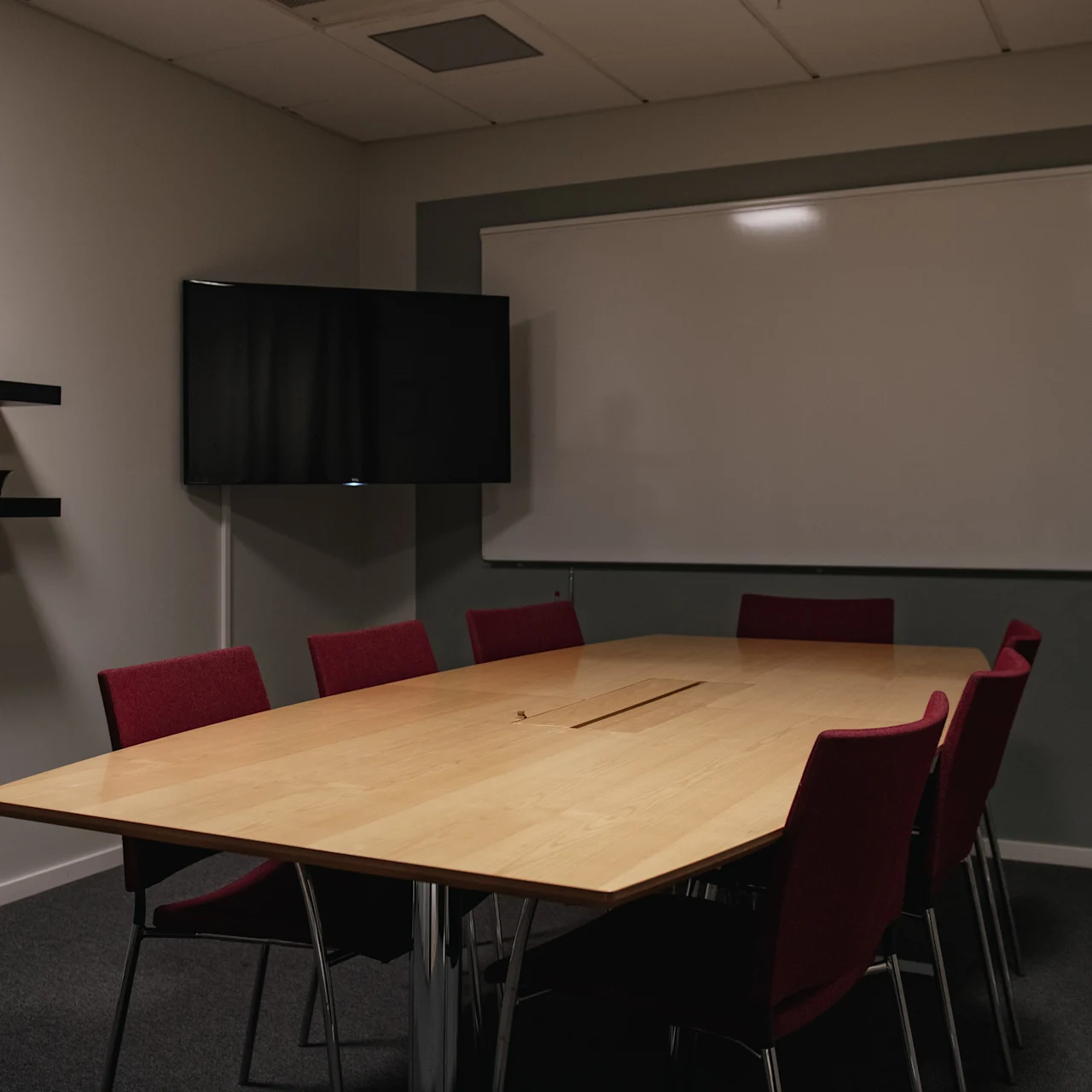 A wooden conference table with red chairs is placed in a meeting room. A whiteboard and shelves with plants and a clock decorate the gray walls.