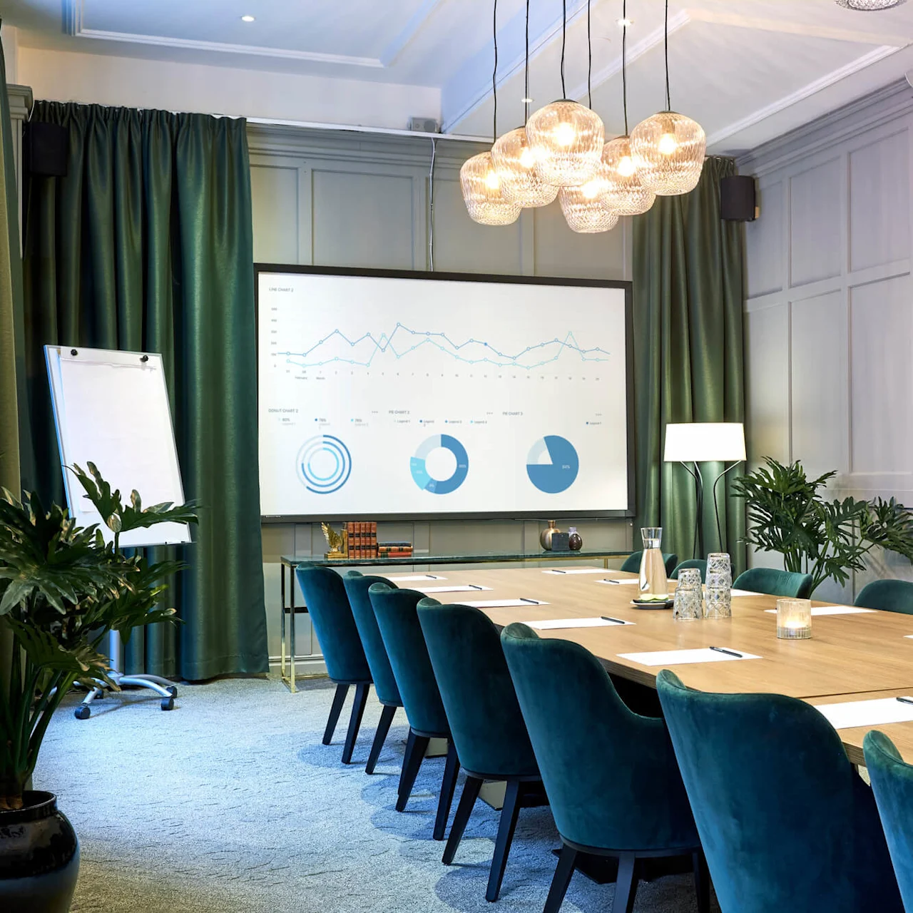 A modern conference room with a presentation screen, a flipchart, green chairs, and a long table set with papers and water.