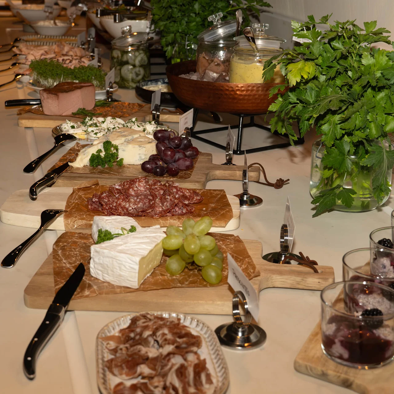 Cheese and charcuterie boards with assorted cheeses, meats, crackers, and grapes are arranged on a buffet table. Nearby, glasses hold desserts topped with blackberries, and fresh herbs in a vase.
