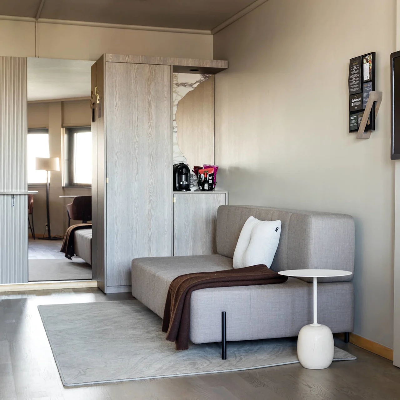 A grey sofa with pillows and a blanket sits beside a small white table. Nearby, a wall-mounted TV and a velvet chair complete the elegant, modern room.