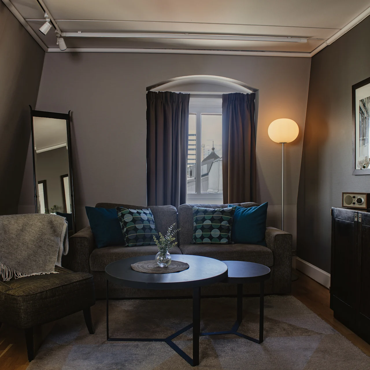 A cozy living room features a grey sofa with blue cushions and a side chair, complemented by a round coffee table. A floor lamp and framed pictures enhance the warm, inviting ambiance.