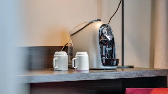 A sleek coffee machine sits on a dark wooden countertop with two white mugs beside it. A floor lamp and beige wall create a cozy ambiance.