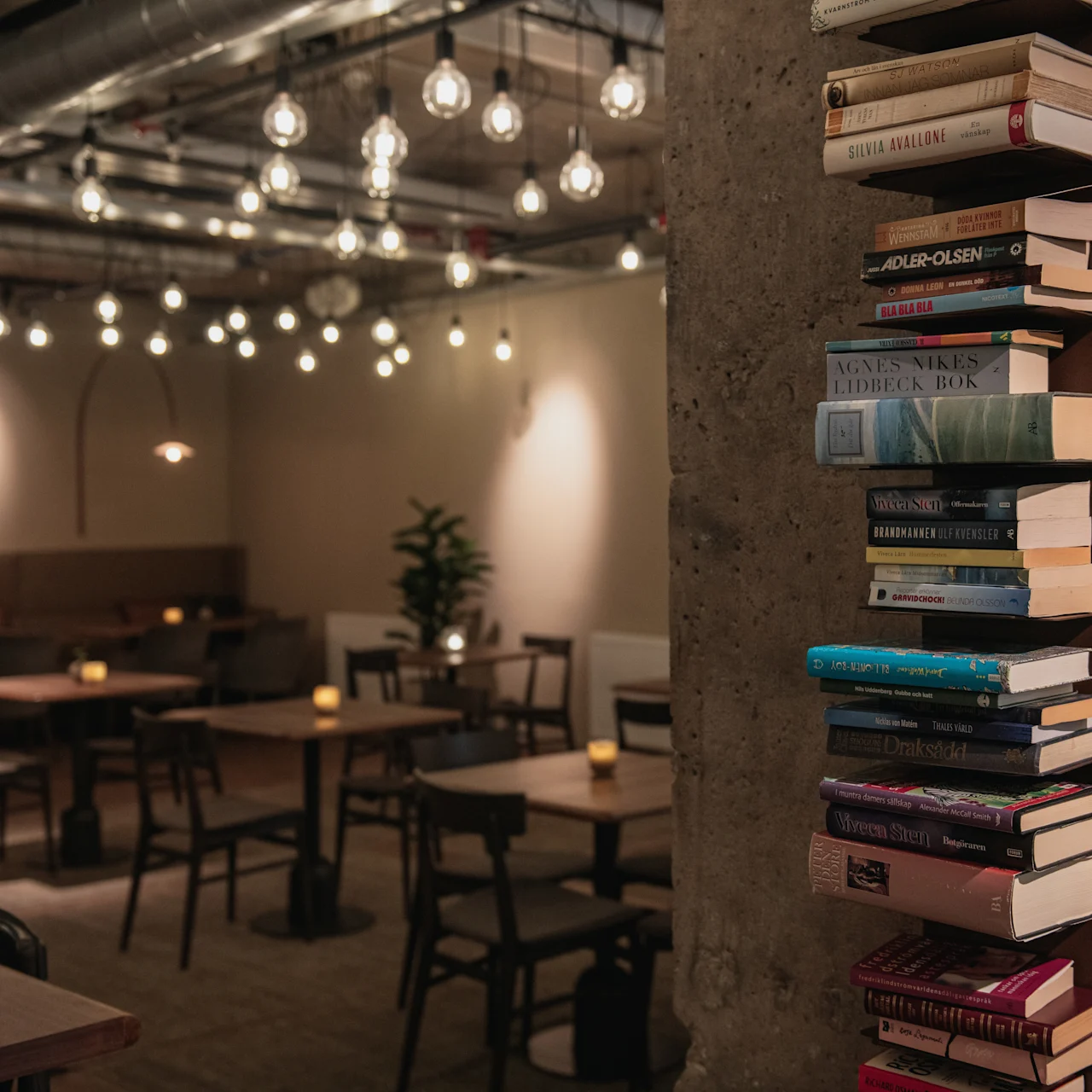 A stack of books is mounted on a concrete column in a dimly lit café. Tables and chairs fill the room, illuminated by hanging bulbs.