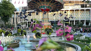 A vibrant amusement park scene with a swing carousel in motion, surrounded by fountains, lush greenery, and colorful flowers.