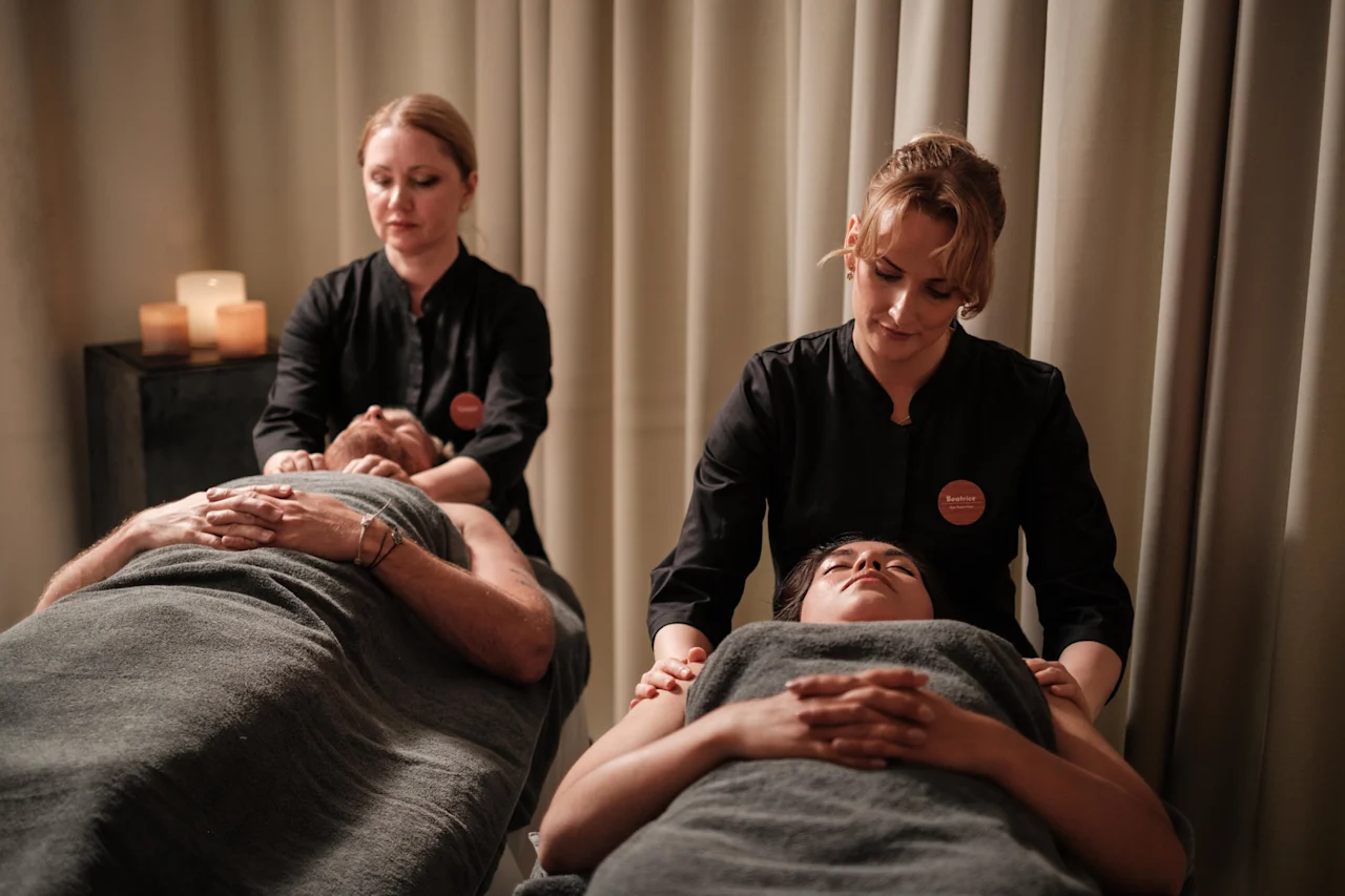Två personer ligger under grå filtar och får massage av massageterapeuter i ett svagt upplyst rum med ljus och skapar en avkopplande atmosfär.