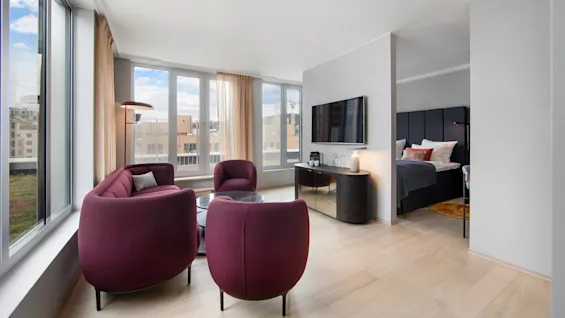Elegant seating area featuring dark red chairs around a small table, adjacent to large windows with city views; a bedroom with a black bed is visible in the background.