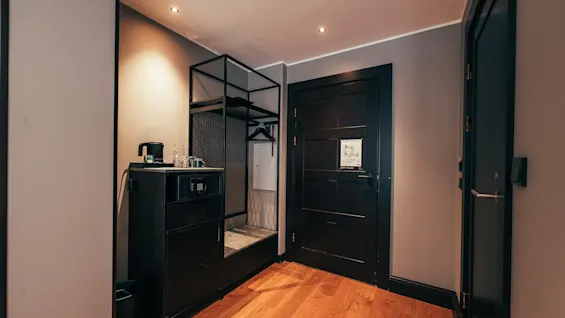 A welcoming hotel room entrance featuring a wooden floor, a convenient cabinet with a kettle, a clothes rack, and two dark doors.