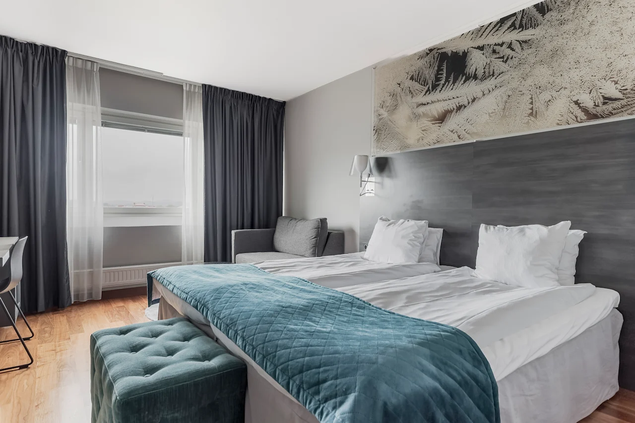 A comfortable hotel room with two beds, a cozy blue throw, and a window offering natural light. Perfect for relaxation.