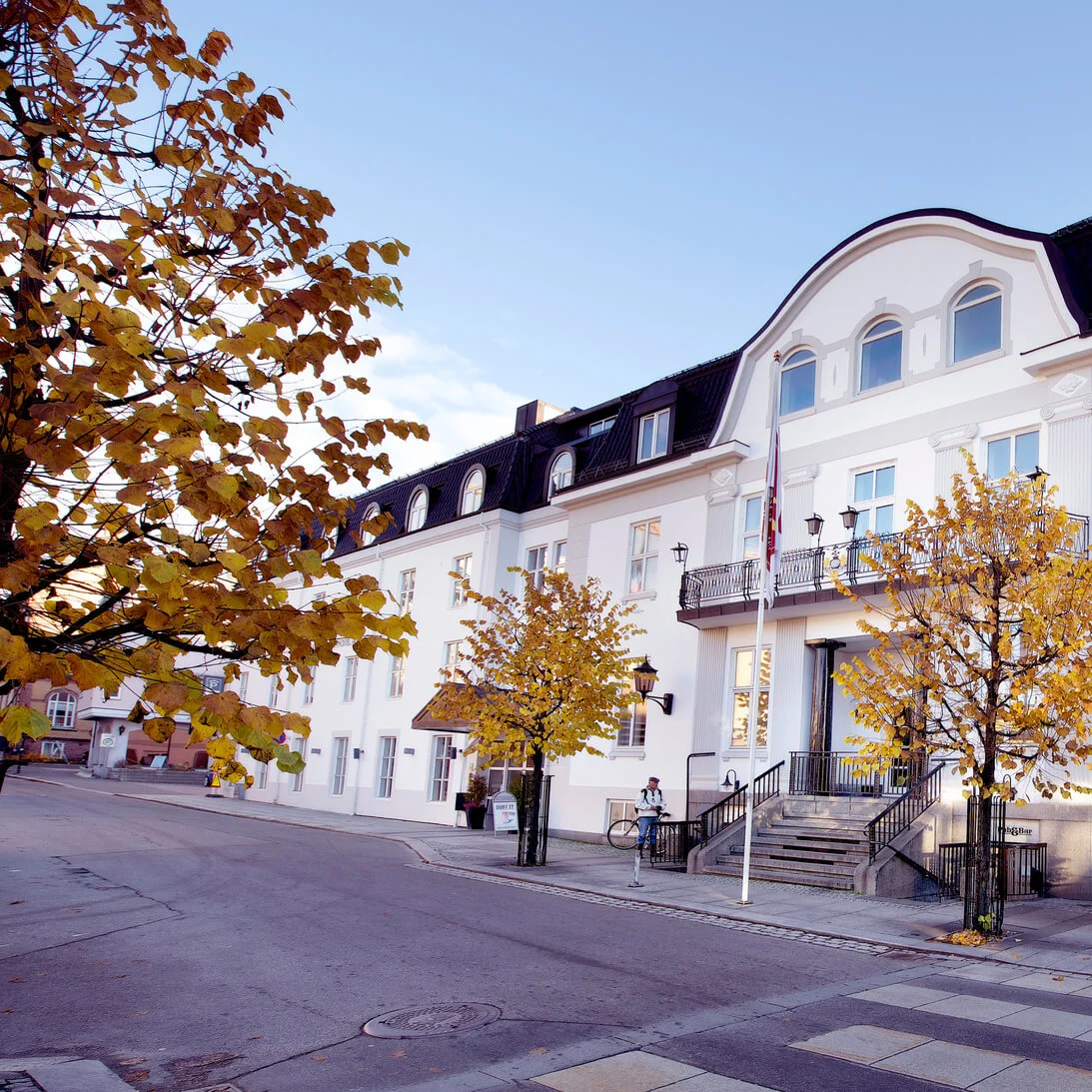En vit trestjärnig byggnad med svart tak, balkonger och höstträd i en stadsinställning under dagtid.