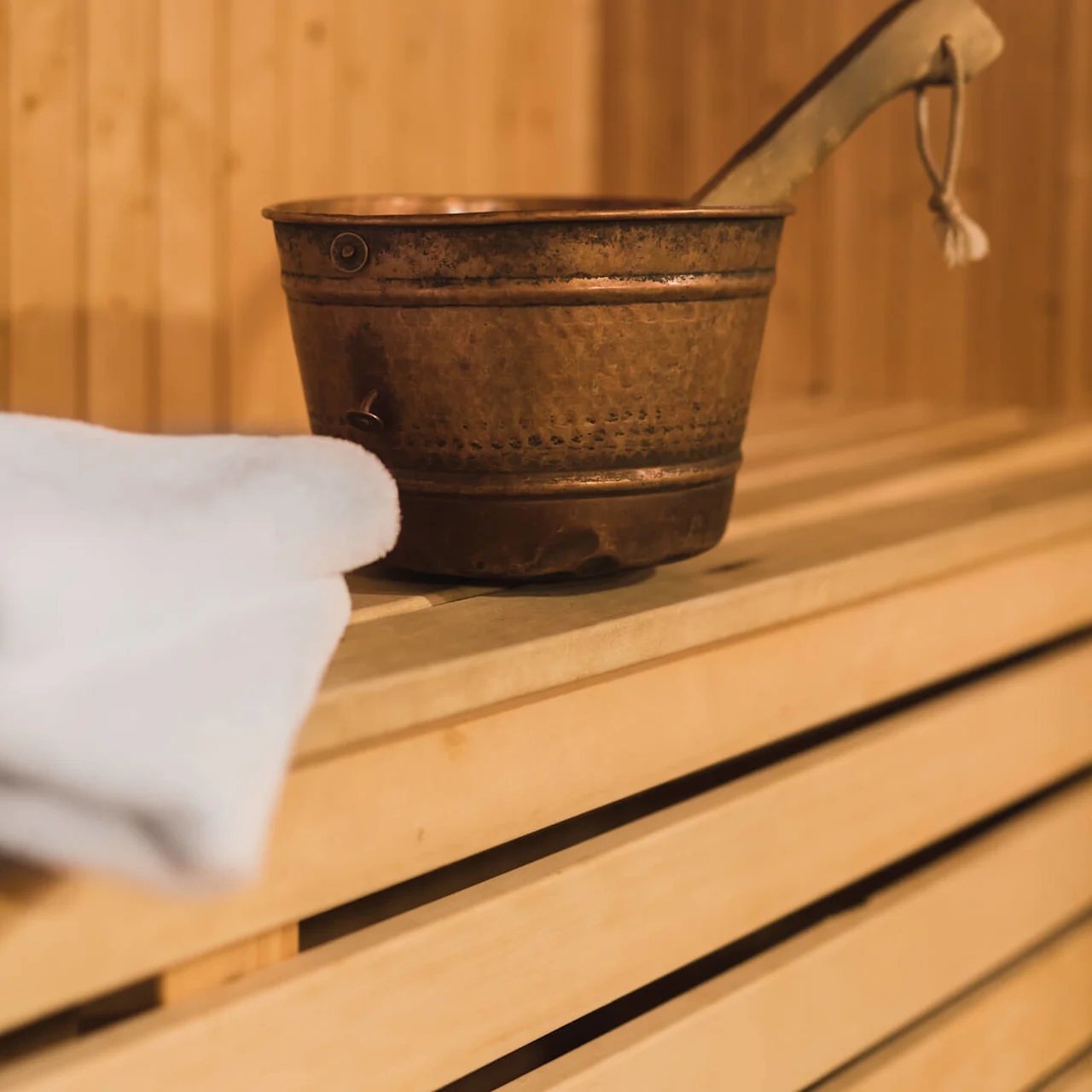 A copper sauna bucket with water ladle, resting on wooden sauna benches, with a white towel nearby.