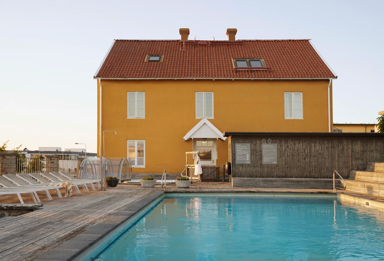 En livlig gul byggnad med rött tak vetter mot en uppfriskande utomhuspool med solstolar en solig dag.