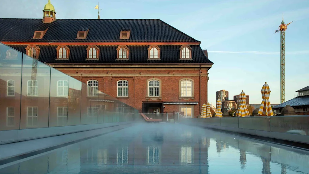 En tåget pool reflekterer en historisk murstensbygning med kvistevinduer, omgivet af lukkede gulstribede parasoller i en bymæssig sammenhæng med et højt, dekorativt tårn i baggrunden.