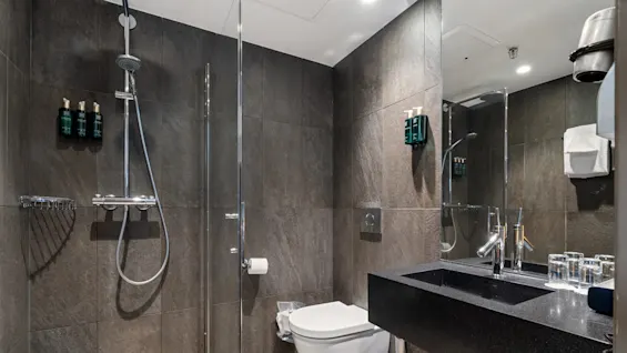 Et moderne badeværelse med en brusekabine med glasvægge, toiletartikler, en vægmonteret toilet og en sort bordpladeservant. Mørke fliser dækker væggene, suppleret med en række glas og en hårtørrer.