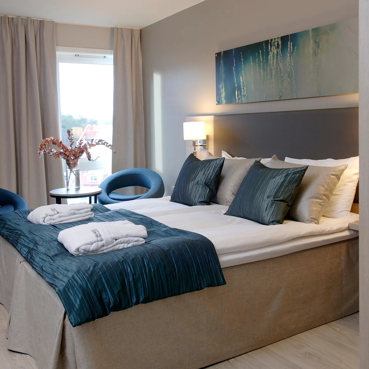 En seng med teal-fargede puter omgitt av moderne møbler og kunst. Beige gardiner rammer inn et vindu, og morgenkåper ligger brettet på sengen.