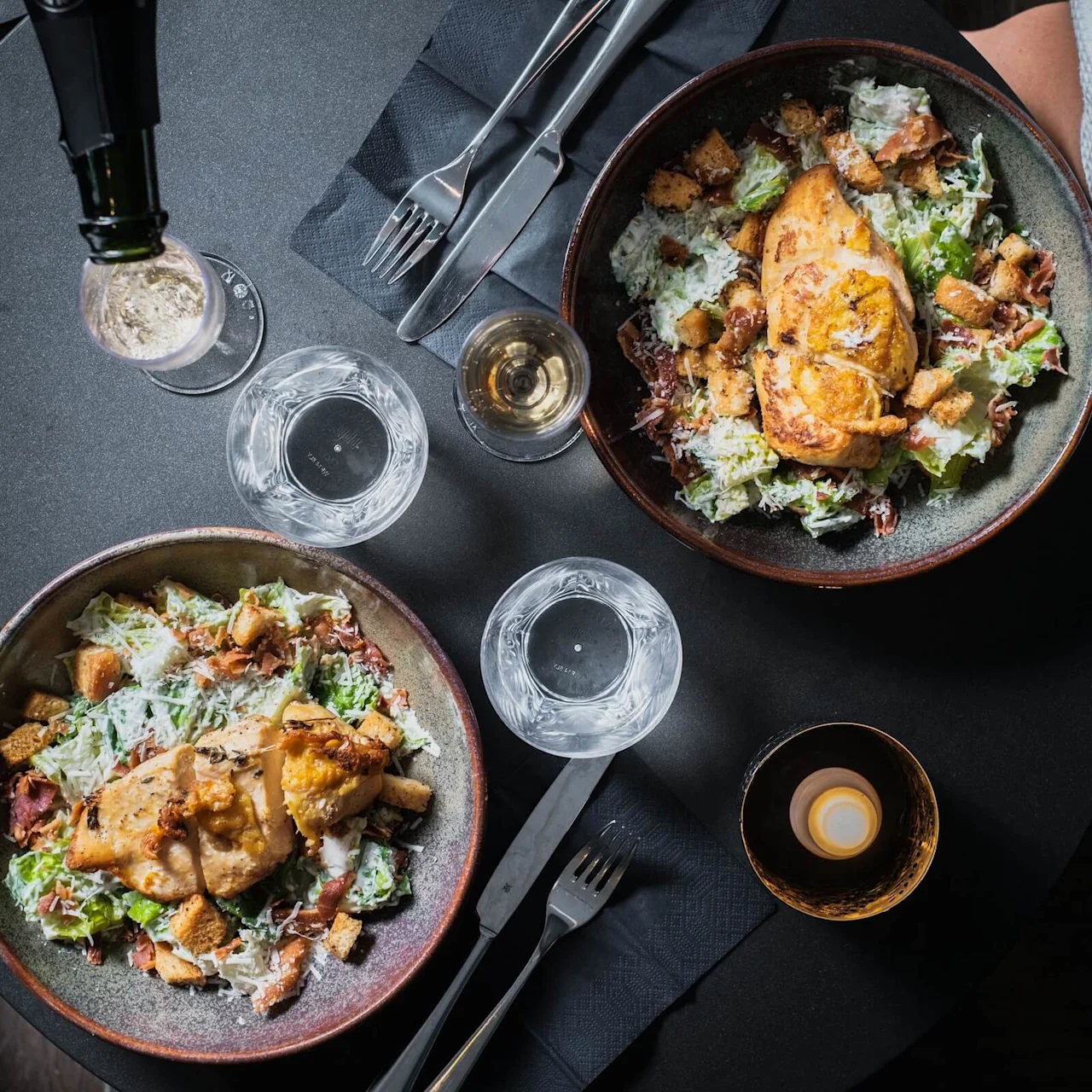 Two bowls of Caesar salad with grilled chicken are arranged on a dark table, accompanied by glasses of water, a glass of white wine, and a lit candle.