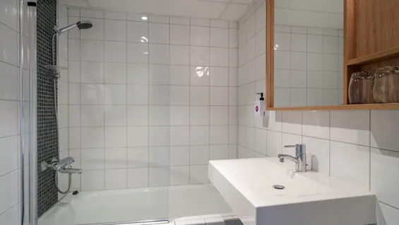 A bathtub with a glass shower screen and showerhead is on the left. A rectangular sink with a chrome faucet is beside it. The walls are tiled white.