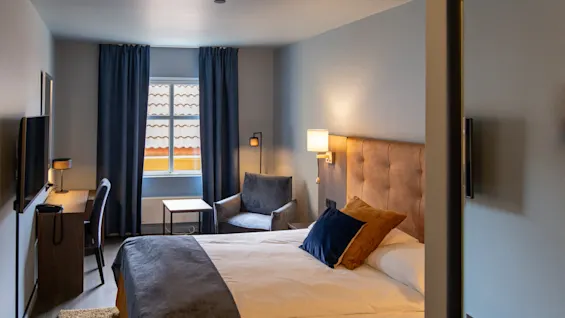 Bed rests under a window with blue curtains, featuring a headboard and cushions. A chair and desk are nearby. Warm lighting enhances the cozy room, with no visible text.