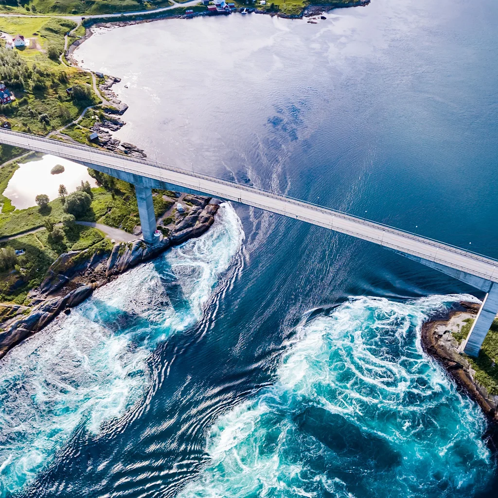 Flygfoto över en bro som sträcker sig över ett sund med kraftiga turkosa strömmar och virvlar, omgiven av grönt landskap.