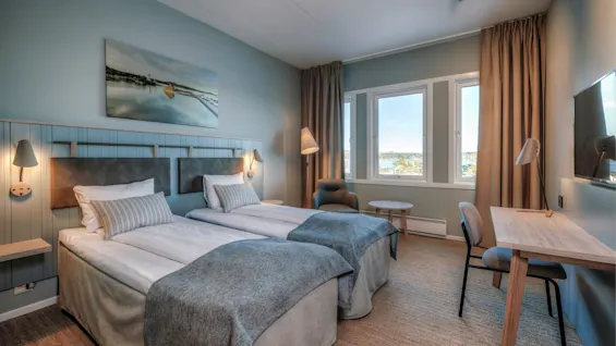 Twin beds with pillows and blankets stand against a light blue wall adorned with a serene landscape painting. A window provides a view of a distant landscape, complemented by a desk and chair.
