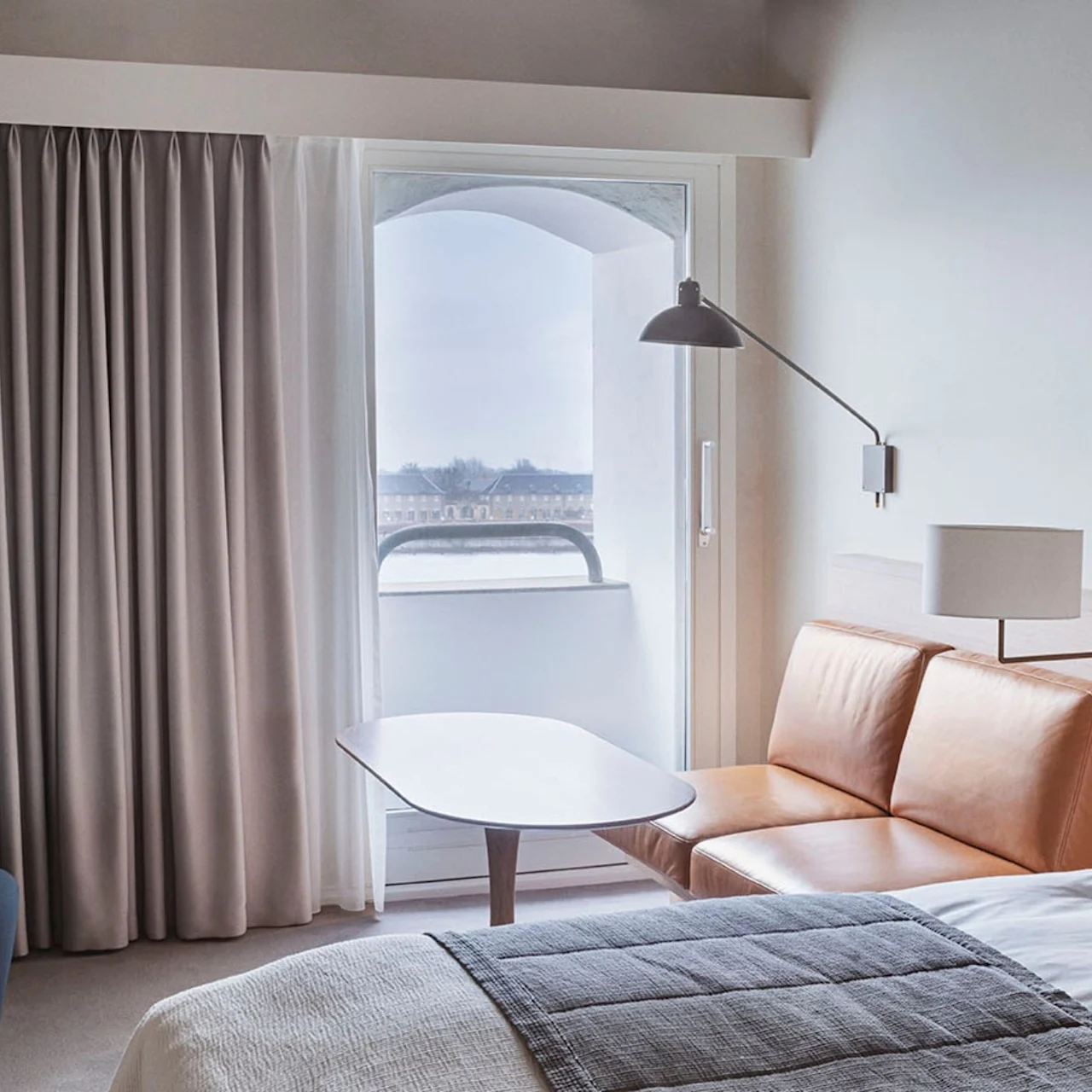 A cozy hotel room features a brown leather sofa near a window with sheer curtains. A small lamp hangs above, and a neatly made bed is visible. A soft armchair sits nearby.