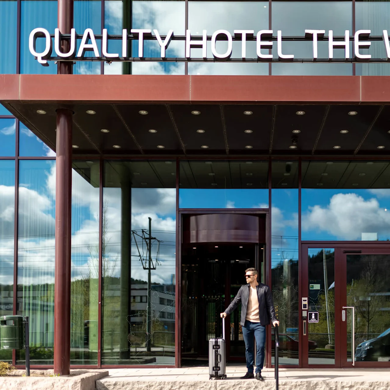 En man med en resväska lämnar ett hotell med glasfasad, under en skylt där det står "QUALITY HOTEL THE WEAVER" en solig dag som reflekteras i fönstren.