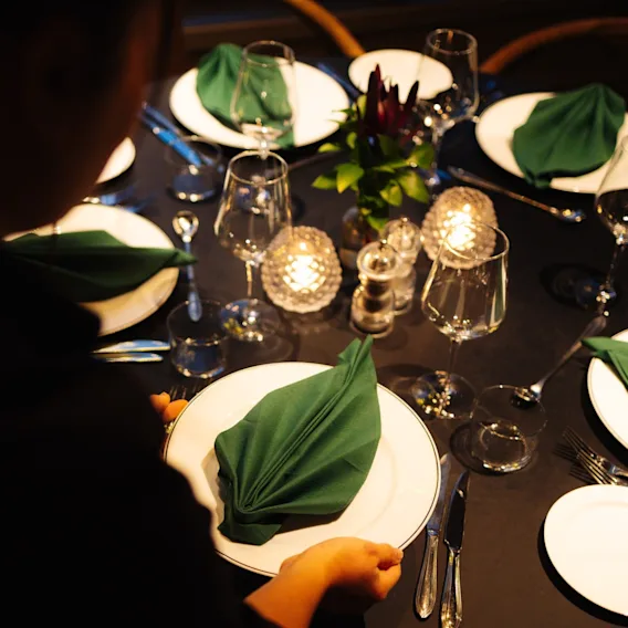 En person dekker et vakkert arrangert middagsbord med hvite tallerkener, grønne servietter, vinglass og tente lys.