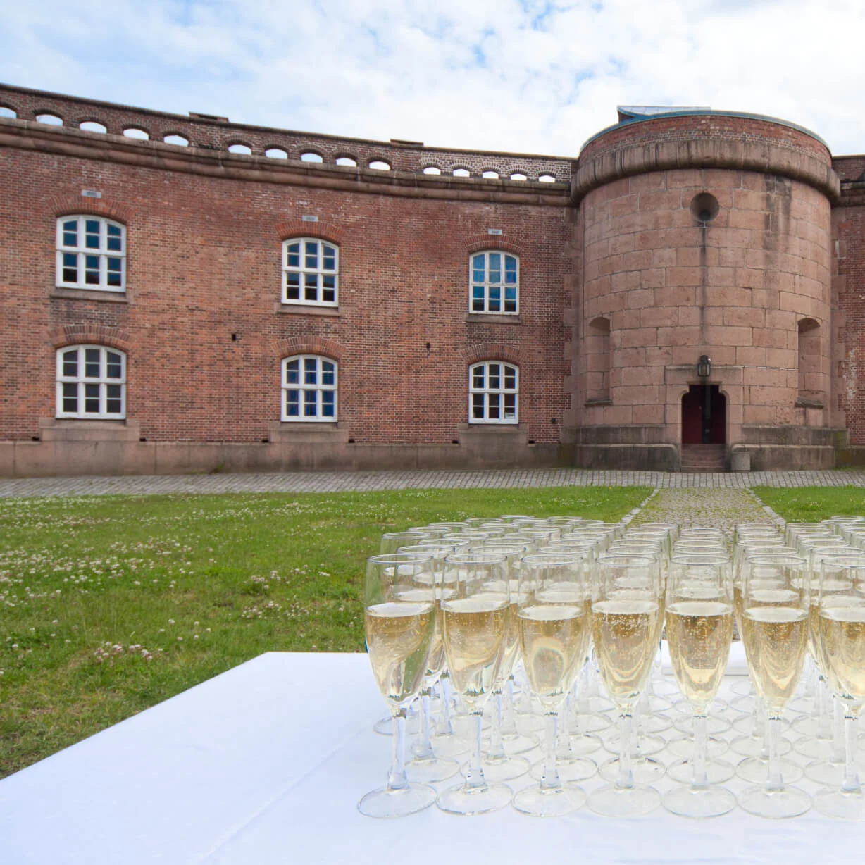 Champagneglass arrangert på et hvitt bord foran en historisk murbygning med buede vinduer. Miljøet er utendørs på et grønt område.